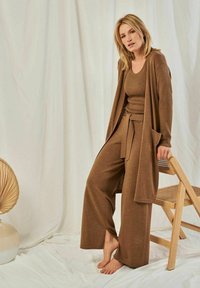 Braunes geripptes Strick-Dreiteiler-Loungewear-Set mit einem langen Cardigan, einem engen Oberteil und weit geschnittenen Hosen. Holzstuhl und dekorative Vase im Hintergrund.