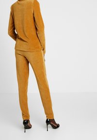 Corduroy loungewear sæt i sennepsgul, der består af en langærmet top og matchende bukser med ribbet tekstur og tætsiddende silhuet. Sorte højhælede sko.