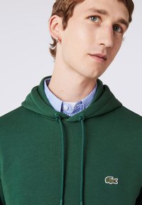 Jeune homme portant un sweat à capuche vert sur une chemise à carreaux bleue avec un petit logo crocodile sur la poitrine et une boucle d’oreille anneau.