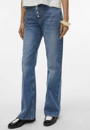 Jeans en denim bleu clair évasés avec une taille haute, fermeture à cinq boutons à l'avant et coutures visibles. Portés avec des chaussures noires ayant un accent brillant.