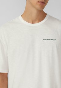 Witte T-shirt met korte mouwen, gemaakt van zachte stof, met een ronde hals en een kleine groene geborduurde tekst "Abonneer op de Natuur" op de borst.