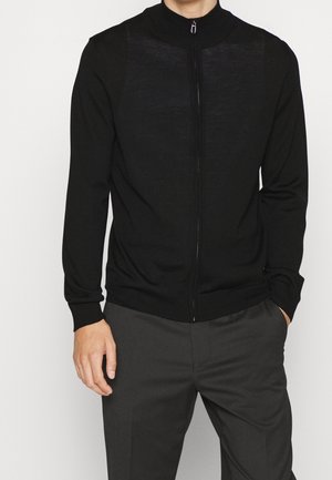 Homme portant un pull noir zippé et un pantalon gris foncé, debout contre un fond blanc uni, les mains détendues le long du corps.