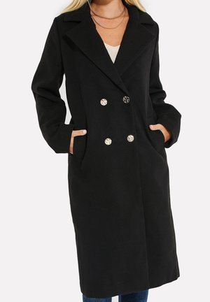 Frau trägt einen langen schwarzen, zweireihigen Mantel mit Tiger-Print-Knöpfen, Hände in den Taschen und ein weißes Oberteil darunter.