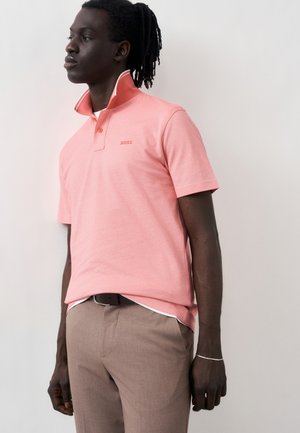 Άνδρας που φοράει κοραλλί ροζ πόλο με όρθιο γιακά και μπεζ παντελόνι, στέκεται μπροστά σε απλό ανοιχτό φόντο, κοιτάζοντας στο πλάι.