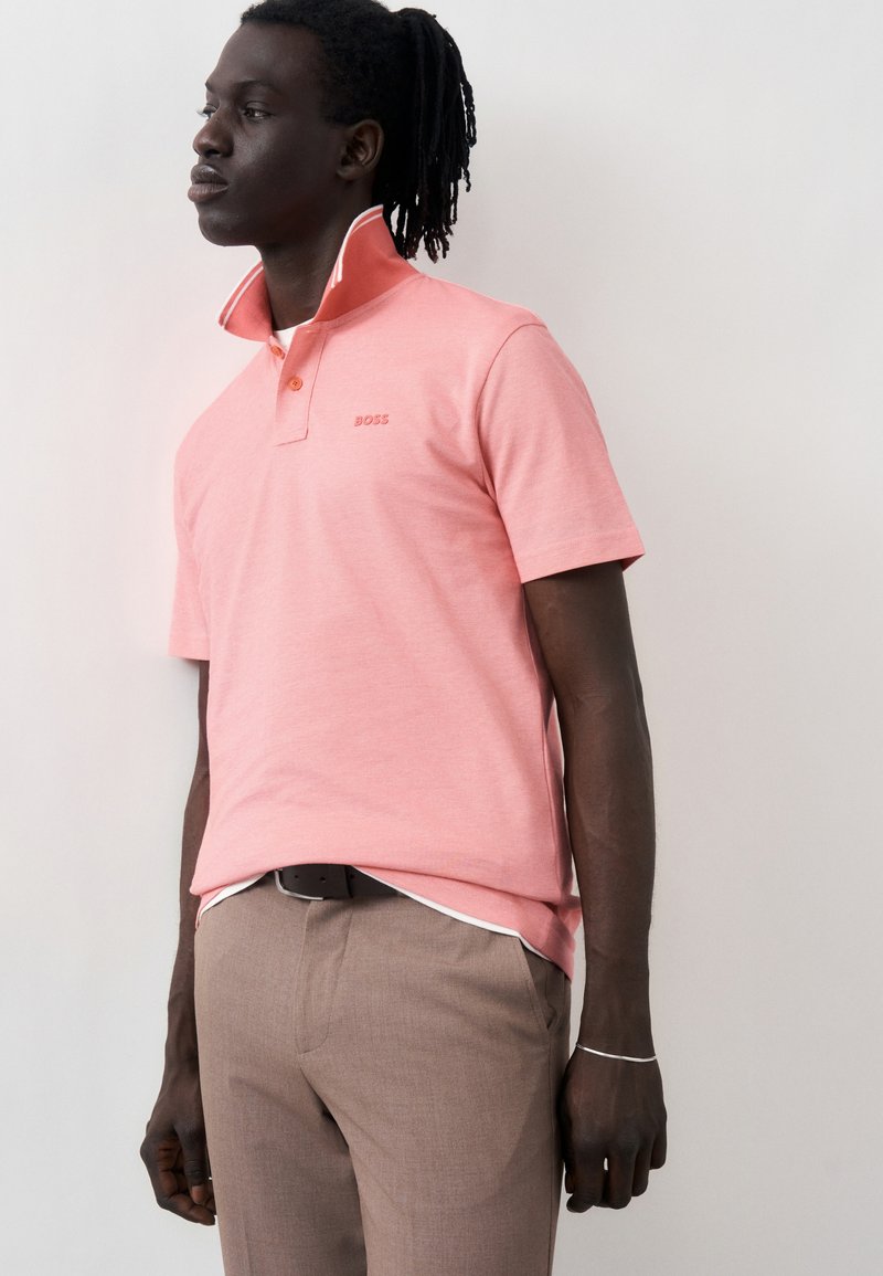 Άνδρας που φοράει κοραλλί ροζ πόλο με όρθιο γιακά και μπεζ παντελόνι, στέκεται μπροστά σε απλό ανοιχτό φόντο, κοιτάζοντας στο πλάι.