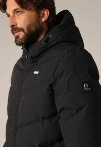 Veste matelassée noire avec capuche, présentant des lignes matelassées, une fermeture éclair frontale et un écusson de marque sur la manche. Tissu lisse et résistant aux intempéries.