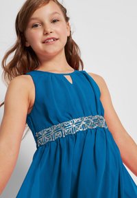 Abito senza maniche blu con gonna plissè, dotato di una cintura decorativa con paillettes argento. Scollo a chiave e texture del tessuto liscia.