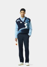 Marineblauer Strickpullunder mit weißem und blauem Wellenmuster über einem hellblauen Hemd mit Knöpfen, kombiniert mit dunklen Hosen und weißen Sneakers, während eine Blume gehalten wird.