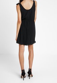 Robe noire sans manches avec une taille cintrée, une jupe fluide et un nœud dans le dos. Associée à des sandales à talons hauts noires avec des découpes.