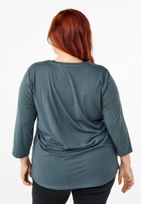Dark blue, long-sleeve top featuring a round neckline and a loose fit. Smooth texture with subtle draping at the back. Black pants visible.