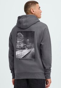 Man in een grijze hoodie met een zwart-wit stadslandschapprint en de tekst "BLEND SINCE 1993" op de achterkant, gezien van achteren.