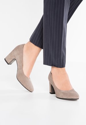 Escarpins en daim beige avec un bout arrondi et un talon bloc. Portés avec un pantalon bleu marine à rayures fines, mettant en valeur une texture lisse.