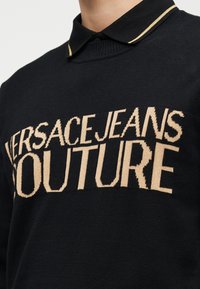 Μαύρο πουλόβερ με μπεζ κεντημένο κείμενο "VERSACE JEANS COUTURE" κατά μήκος του στήθους, φορεμένο πάνω από μαύρο πουκάμισο με γιακά και χρυσή επένδυση.