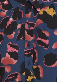 Multikleurige blouse met een marineblauwe achtergrond, voorzien van roze en zwarte abstracte vormen. Het heeft een kraag en blauwe knopen langs de voorkant.