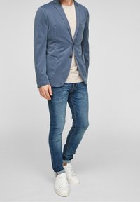 Jasnoniebieska bawełniana marynarka z klapkami, z przednimi kieszeniami; noszona na beżowej koszulce, w zestawieniu z dopasowanymi niebieskimi dżinsami i białymi adidasami.