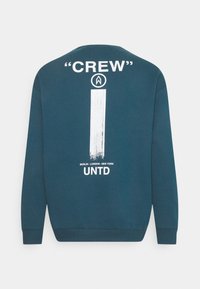 Sudadera azul verdoso con cuello redondo y mangas largas. Los gráficos en blanco incluyen "CREW," una pincelada vertical y el texto "BERLÍN - LONDRES - NUEVA YORK."