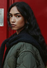 Gepatchte olivgrüne Jacke mit einem schwarzen Schal. Lange, wellige, dunkle Haare und dezentes Make-up vor einem roten Hintergrund.
