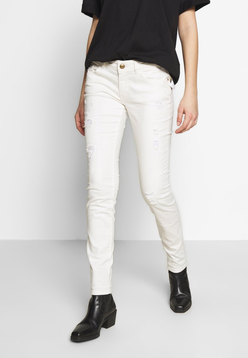 Jeans skinny blancos con parches desgastados, diseño de cinco bolsillos y un botón dorado. Combinados con botas negras hasta el tobillo.