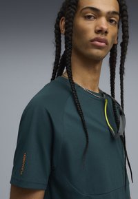 Junge Person mit langen, geflochtenen Haaren, die ein dunkelgrünes PUMATECH-Shirt und eine Sonnenbrille, die am Hals hängt, vor einem grauen Hintergrund trägt.
