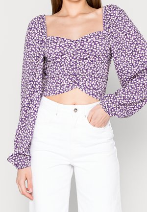 Femme portant un chemisier violet à manches longues et court avec un imprimé floral blanc et un pantalon blanc taille haute, la main dans la poche.