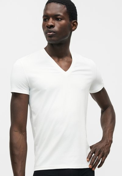 Νέος άνδρας που φοράει απλό λευκό T-shirt με V-λαιμόκοψη και κοντά μανίκια, στέκεται με το ένα χέρι στη μέση μπροστά από λευκό φόντο.