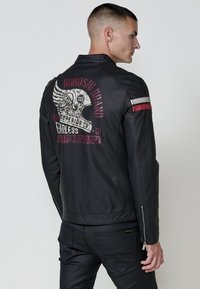 Uomo in giacca di pelle nera con grafica di un casco alato e testo rosso sul retro, in piedi di lato su uno sfondo semplice.