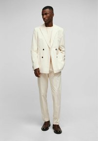 Helle, zweireihige Anzugjacke mit strukturiertem Finish, kombiniert mit passenden Hosen. Schwarze Schuhe runden das Ensemble ab.