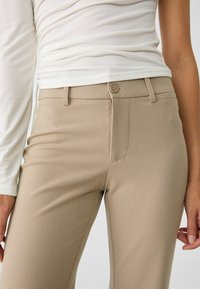 Vrouw in beige op maat gemaakte broek en een wit lange mouwen top, staande met haar handen ontspannen naast zich.