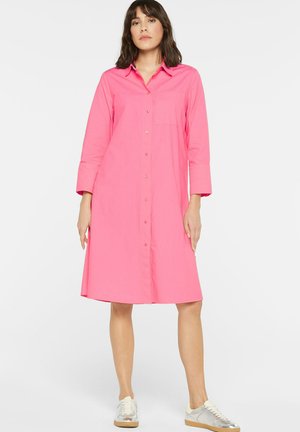Woman standing wearing a knee-length bright pink button-up shirt dress with long sleeves and white sneakers.