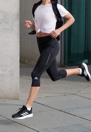 Person, der løber udendørs iført sorte Adidas korte leggings, sorte sneakers med hvide striber, hvid knyttet skjorte og sort ærmeløs jakke.