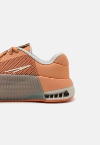 Zbliżenie na pomarańczowy i szary but sportowy, koncentrując się na siateczkowej cholewce, wyściełanym pięcie oraz wentylowanej konstrukcji podeszwy środkowej.