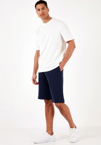 T-shirt bianca in cotone abbinata a pantaloni corti blu navy, con tasche laterali. Il modello indossa sneakers sportive bianche, in una posa rilassata.
