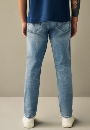 Lichtblauwe denim jeans met een rechte pasvorm, voorzien van details op de achterzakken. Witte sneakers zichtbaar bij de enkels. Eenvoudig ontwerp, zonder patronen.