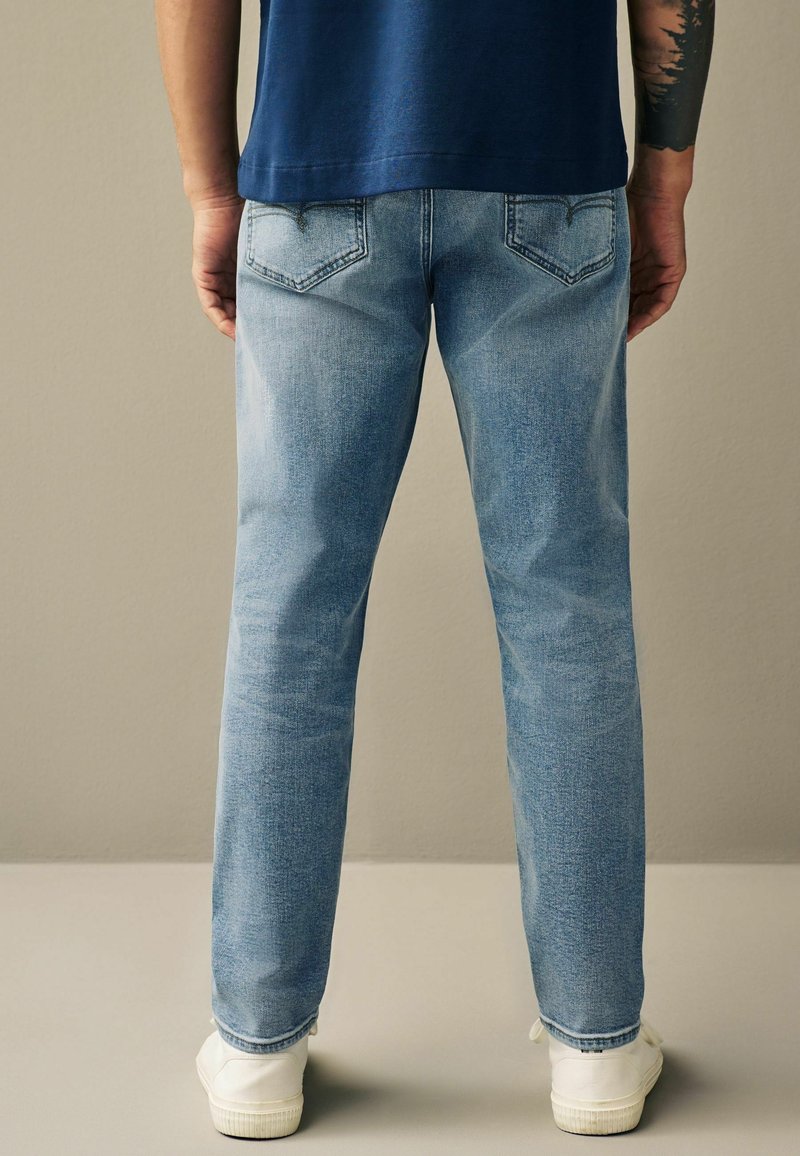 Jean en denim bleu clair à coupe droite, avec des détails sur les poches arrière. Baskets blanches visibles aux chevilles. Design simple, sans motifs.