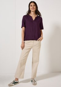 Blusa viola a maniche corte con scollatura a V, abbinata a pantaloni beige a gamba larga. L'outfit è completato da sneaker grigie e verdi casual.