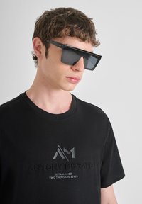 Crna majica kratkih rukava s podignutim dizajnom loga u sredini, u kombinaciji s velikim crnim pravokutnim naočalama s metalnim detaljima.