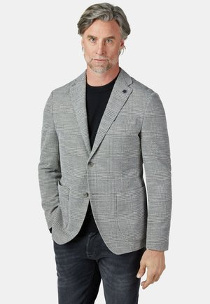 Mann mit grauem Haar, der ein grau kariertes Sakko über einem schwarzen Hemd und dunklen Jeans trägt, steht mit einer Hand in der Tasche vor weißem Hintergrund.
