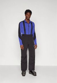 Homem vestido com uma jaqueta zip-up azul e preta, calças de esqui pretas com suspensórios e botas de esqui pretas, em pé contra um fundo simples.