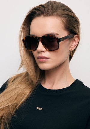 Vrouw met lang blond haar draagt grote schildpadzonnebril, gouden hangende oorbel en zwarte top met klein goud logo.