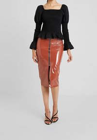 Svart smocktopp med puffärmar, kombinerad med en glansig rostfärgad pennkjol med framdragkedja och slits. Svarta sandaler med remmar.