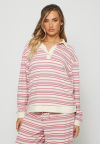 Kobieta z długimi blond włosami nosząca różowo-białą i jasnoniebieską paskowaną koszulę polo z długim rękawem oraz pasujące spodnie na jednolitym tle.