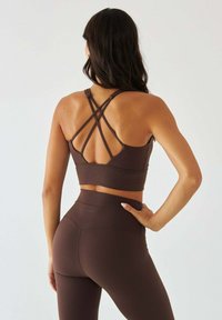 Brązowy sportowy biustonosz z krzyżującym się tyłem, w zestawie z pasującymi do niego legginsami z wysokim stanem. Gładka faktura tkaniny, minimalne detale z logo.