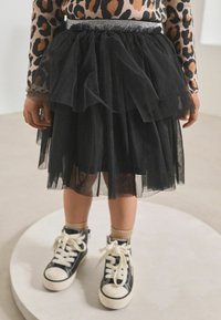 Schwarzer Tüllrock mit gestuften Lagen und einem glitzernden Taillenbund, kombiniert mit schwarzen High-Top-Sneakern und beigen Socken. Texturiertes und voluminöses Design.
