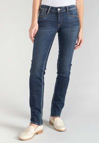 Jeans ajustados de denim en azul oscuro, con bolsillos frontales, costuras visibles y una cintura de altura media. Combinados con mocasines blancos.