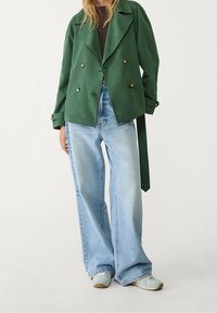 Grøn dobbeltradet jakke med store reverser, parret med lyseblå vidde jeans og lyseblå sneakers. Glat, letvægtsstof.