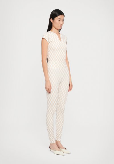 Strakke jumpsuit in perzik met een crème geometrisch patroon, met een V-hals en korte mouwen. Gecombineerd met witte instapschoenen.