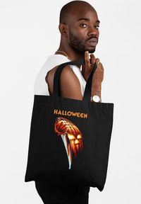 Czarna torba na ramię z graficznym wzorem przedstawiającym nóż i dynię ze świecącymi oczami, z napisem "HALLOWEEN" w pomarańczowym kolorze nad obrazem.