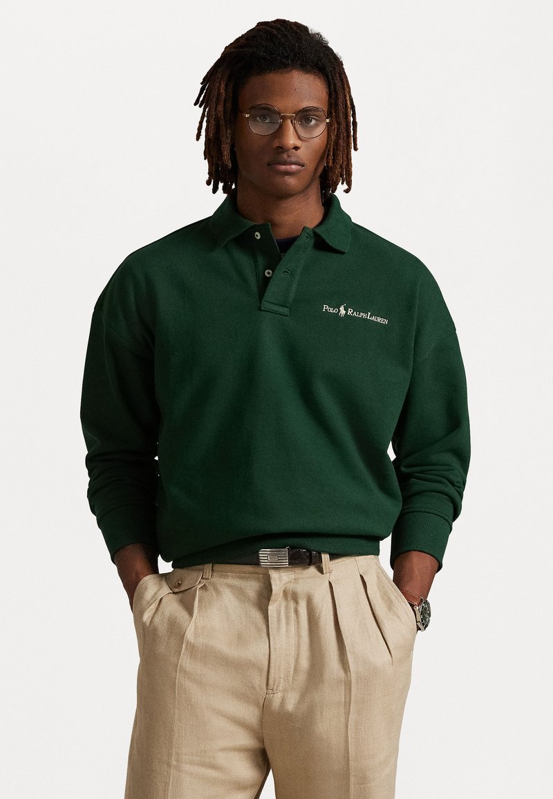 Mørkegrønn polo skjorte med lange ermer, med en logo på brystet. Kombinert med lyse beige plissert bukser og et svart belte.