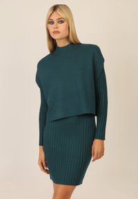 Türkises Strickkleid mit einem verkürzten, weiten Oberteil und einem gerippten Rock. Lange, eng anliegende Ärmel und ein hoher Kragen. Weiche Textur insgesamt.