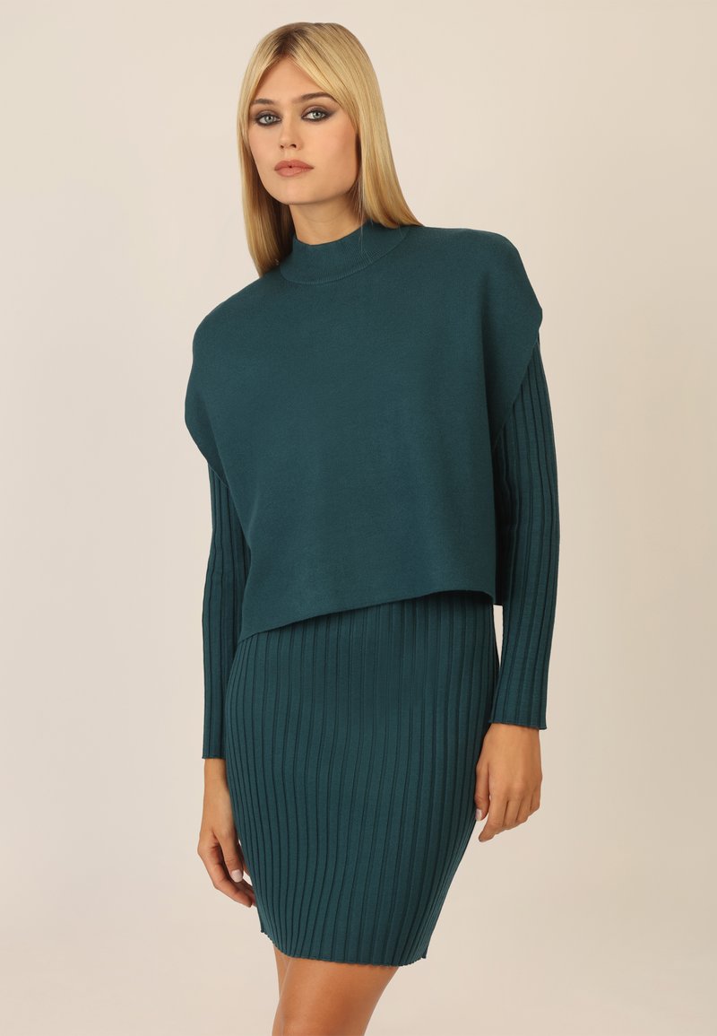 Türkises Strickkleid mit einem verkürzten, weiten Oberteil und einem gerippten Rock. Lange, eng anliegende Ärmel und ein hoher Kragen. Weiche Textur insgesamt.